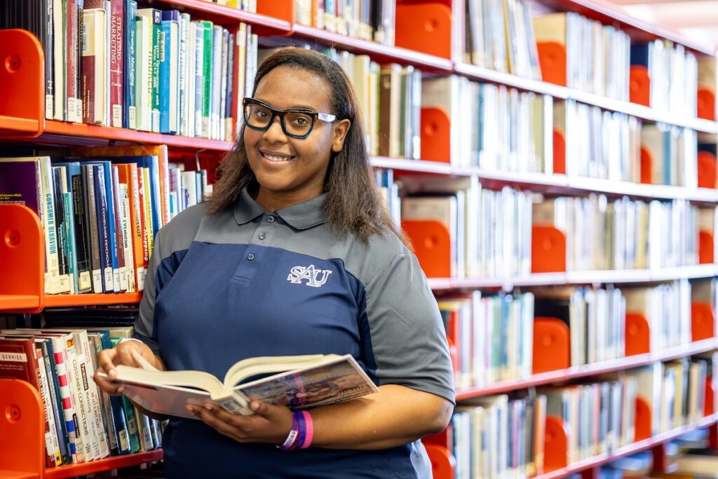 Female student with black rimmed glasses holding an open book, smiling and standing in the library in front of shelves of books